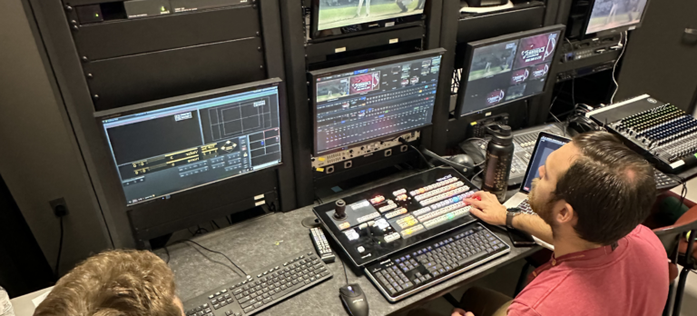 An instructor sits at a bank of TV monitors next to a student.